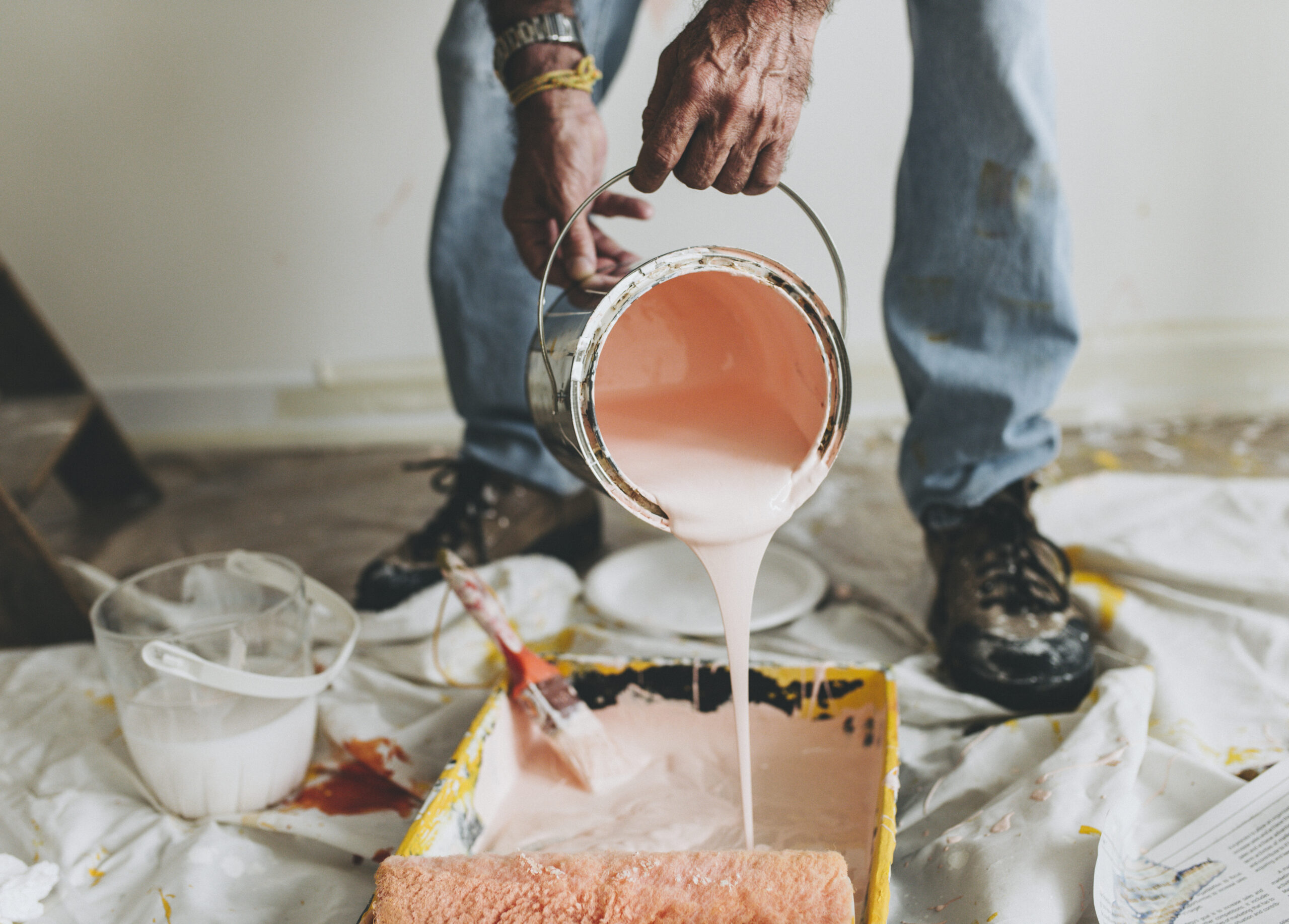 Close-up of a person pouring pink paint from a can into a tray during a painting project, with a paint roller nearby.
